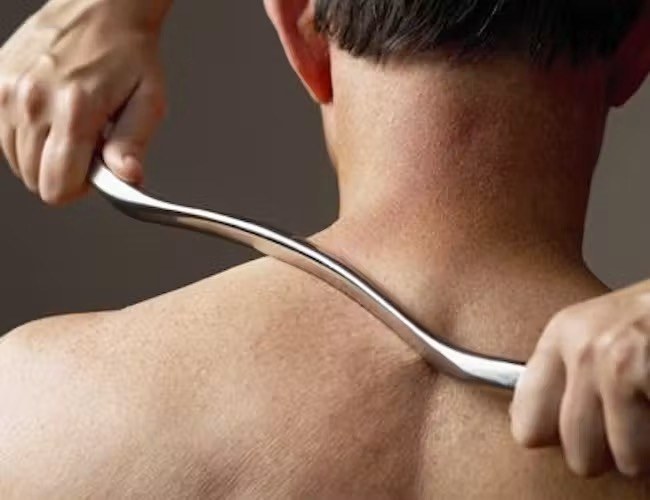 A practitioner drawing a curved steel Graston tool across the top of a patient's shoulder and neck